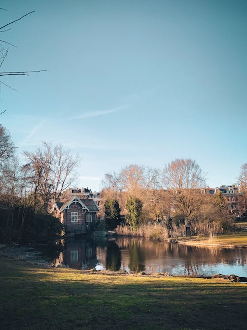 A calm, scenic view of a residential area near a small lake, with a brick house featuring large windows and a sloped roof situated among trees and shrubs on the opposite side of the water. Bare branch