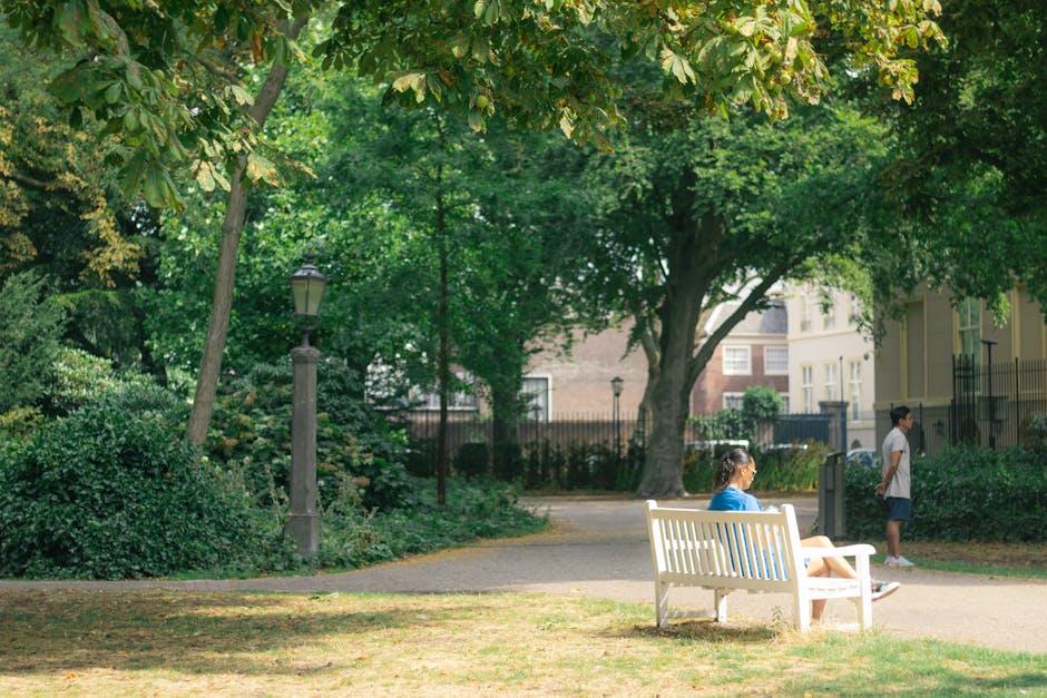 A woman in a blue shirt is sitting on a white wooden park bench reading or resting, while a young girl in a white shirt and dark shorts stands nearby on a paved pathway within a green park. The park f